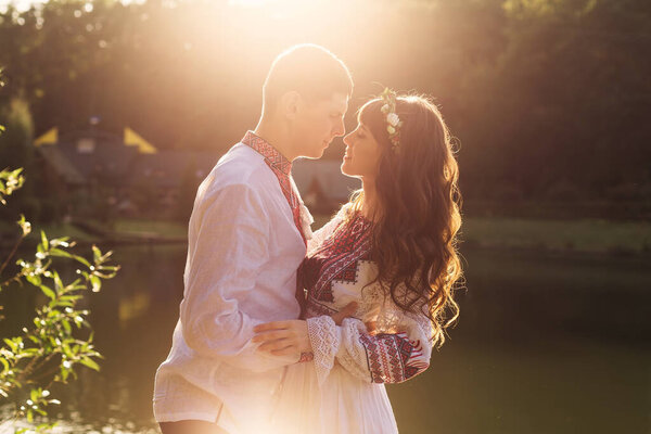 couple in love in embroidered clothes by the lake. profile of guy and girl with closed eyes. sunlight shines on them. 