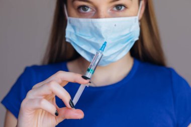 Maskeli bir kadın doktor elinde şırıngayla arka planda bulanık bir kadının şırıngasını tutuyor.