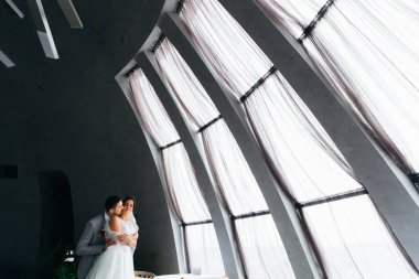 Damat gelini arkadan kucaklıyor büyük panoramik pencerelerin yanında. Restoran salonu