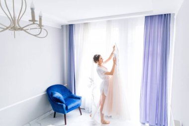 Bornozlu ve duvak takmış gelin pencerenin kenarında duruyor ve gelinlikli bir askı tutuyor..