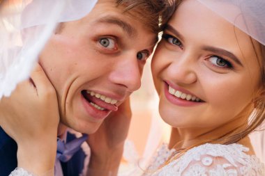 Yeni evli çiftlerin yüzlerini duvak altında kapatıyoruz. Damat dilini gösteriyor ve gelin gülümsüyor. Kameraya bak..