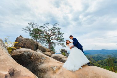 Geriye doğru eğildi ve damat, dağların arka planında kayaların üzerinde ona sarıldı.
