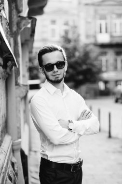 Güneş gözlüklü genç adam kameraya bakar ve siyah beyaz bir fotoğrafta çapraz kollar oluşturur..