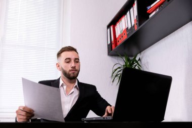 Genç iş adamı dizüstü bilgisayar kullanıyor ve elinde kağıt tutuyor.