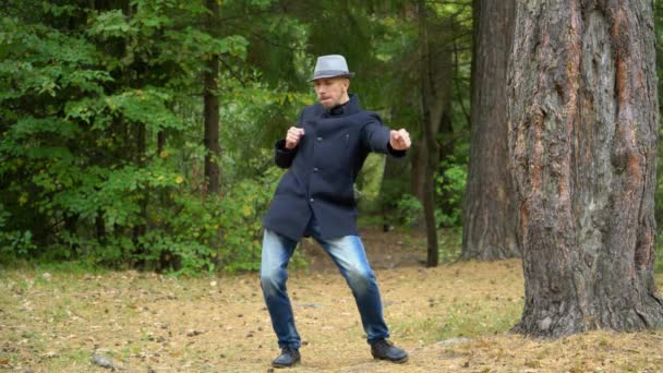 Un hombre de negocios con sombrero y abrigo en el bosque se mueve felizmente — Vídeo de stock