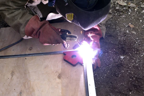 Een sterke man is een lasser in een zwart T-shirt, een masker lassen en lassers leer, een metalen product is gelast met een lastoestel in de garage, blauwe vonken vliegen aan de zijkanten — Stockfoto