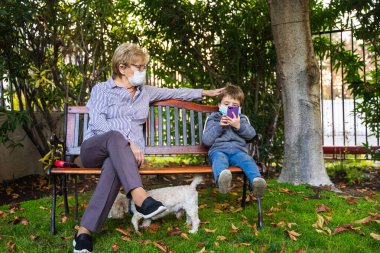 Büyükanne ve küçük çocuk, koronavirüs salgını nedeniyle koruyucu maskeler takıp arka bahçede akıllı telefonla oynuyorlar.