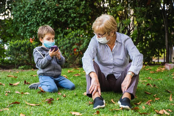 할머니와 아이가 코로나 바이러스로 마스크를 착용하고 뒷마당에서 스마트폰으로 스톡 사진