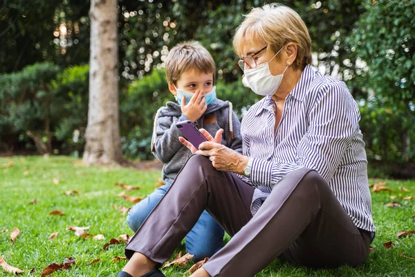 祖母と小さな子供は 保護マスクを身に着けている間に裏庭でスマートフォンで遊んでいるコロナウイルスの流行による ロイヤリティフリーのストック画像