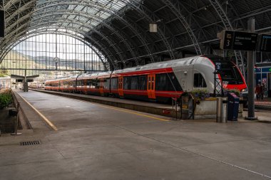 Tren istasyonunda bekliyor. Arka planda dağ manzarası var.