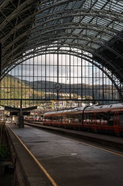 Tren istasyonunda bekliyor. Arka planda dağ manzarası var.