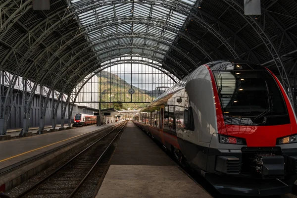 Tren istasyonunda bekliyor. Arka planda dağ manzarası var.
