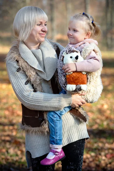 Mom and daughter child with ponytails with soft toy hedgehog smiling in sunny autumn park outdoors