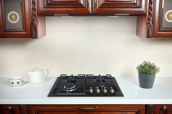 Interior of modern kitchen in classic style with golden elements cherry alder wood cabinetry with built-in appliances, gas hob and extractor fan with flower pot, cup, teapot and copyspace.