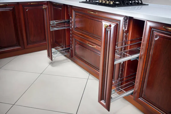Pull out spice rack cabinet filler pantry. Two vertical drawers for spices shelf under countertop with wooden panel. Classic style interior kitchen with golden elements, cherry alder wood cabinetry.
