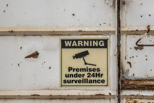 使用されていない工業ビルのドアにある24時間監視警告標識の下で色あせた建物 — ストック写真