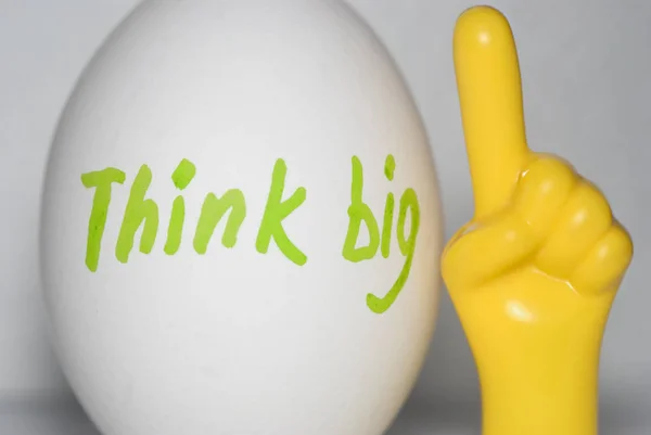 Think big -  the inscription on a white chicken egg, in bright letters in green and a yellow plastic hand with the index finger pointing upwards on a white background. Thinking concept