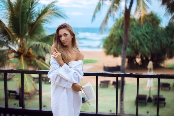 güzel seksi kız balkonda bornoz plaj ve palmiye ağaçları ile ilgili bir kitap ile sabah