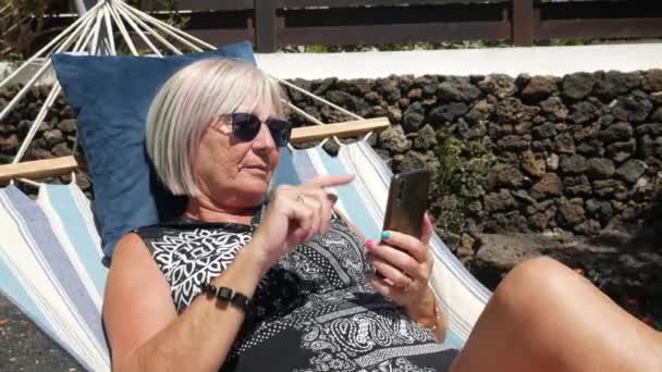 Donna anziana che guarda sul telefono cellulare che si rilassa su un'amaca nel giardino di casa — Video Stock