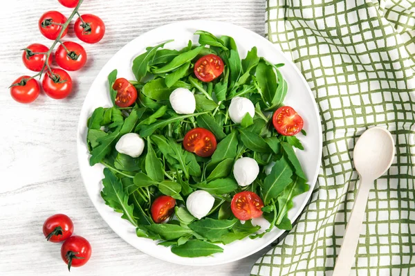 Vegetarian food and healthy lifestyle concept - spring salad with cherry tomato, mozzarella cheese and arugula on a white plate laying on wooden table. Royalty Free Stock Photos