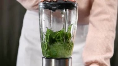 Taze ıspanak yaprakları yavaş çekimde blender içinde öğütülüp suyla karıştırılarak arka planda odaklı olmayan biri ile lezzetli bir smoothie hazırlanıyor. Sağlıklı ve vejetaryen gıda konsepti.