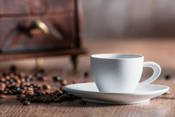 Rustic white porcelain cup with toasted coffee beans and a red mill on the bottom on a wooden table. Relaxation concept and coffee aroma — Stockfoto