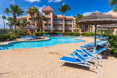 Otel manzarası çok güzel. Açık havuz, mavi güneş yatakları ve yeşil palmiye ağaçlarının arka planında şemsiyeler. Kartal Sahili. Divi köyü golf ve plajı. Oranjestad, Aruba. 09.10.2019.