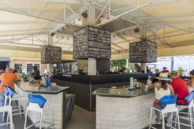 Açık hava barının dışından bak. Barın etrafındaki yüksek mavi beyaz bar sandalyelerinde oturan turistler. Barmen bira ve yemek yapıyor. Oranjestad. Aruba. 09.14.2019