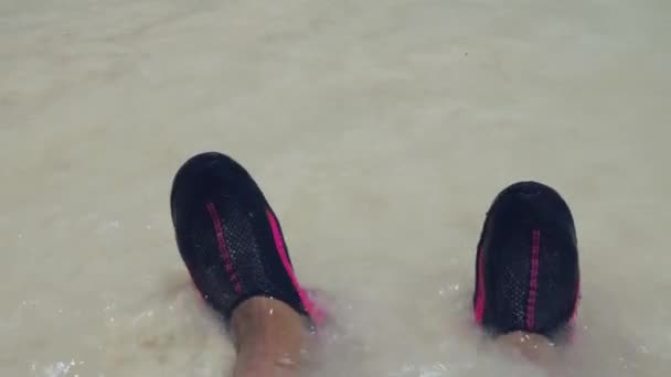水泳シューズで白い砂浜に立っている男性の足で波の圧延のビューを閉じます — ストック動画