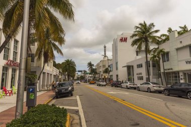 Miami Sahili 'nin güzel manzarası. Asfalt yolun her iki tarafında beyaz binalar ve palmiye ağaçları mavi gökyüzünde ve beyaz bulutlar arka planda. Usa. Miami Güney Sahili. 09.22.2019.