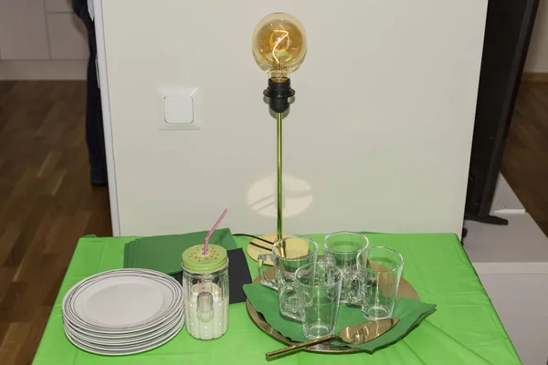 Close up view of lovely small table with white plates and empty glasses prepared for home birthday party. Modern table lamp on green table cloth background.