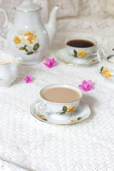 Uma xícara de chá decorada com flores, dois biscoitos e uma vela em uma mesa de madeira branca. No fundo um relógio azul-turquesa em um wainscot amarelo. Hora do chá vintage — Fotografia de Stock