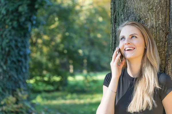 Smějící se atraktivní mladá dívka je telefonování v parku — Stock fotografie