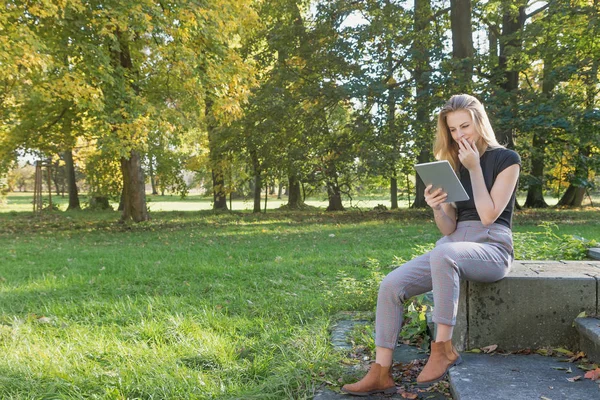 Sonriente jovencita está sentada en el parque mirando la tableta — Foto de Stock