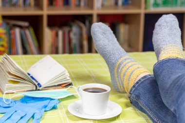 Bacaklar oturma odasında masanın üzerinde. Ayrıca masada defter, kahve fincanı, tek kullanımlık hijyen eldivenleri ve tek kullanımlık koruyucu yüz maskesi var..