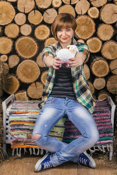 Sevimli genç kısa saçlı kız oturur ve rustik bir bankta oyuncak kuzu tutarak. Ahşap arka plan. Tarım kavramı.