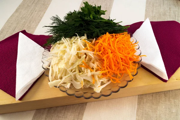 Shredded carrots and cabbage, a bunch of fresh greens - dill and parsley, prepared for salad, against a background of white and claret paper napkins, natural wood and linen cloth.