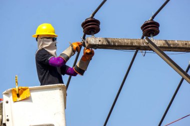 Elektrikçiler açık mavi gökyüzü arka planının altındaki elektrik hattını onarmak için teleferik üzerinde çalışıyorlar..