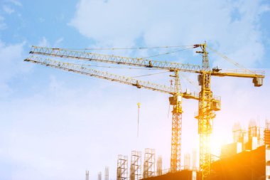 Yükselen vinçler, parlak mavi gökyüzünde inşaat çalışması ve arka planda güneş parlaması olan büyük bulutlar..