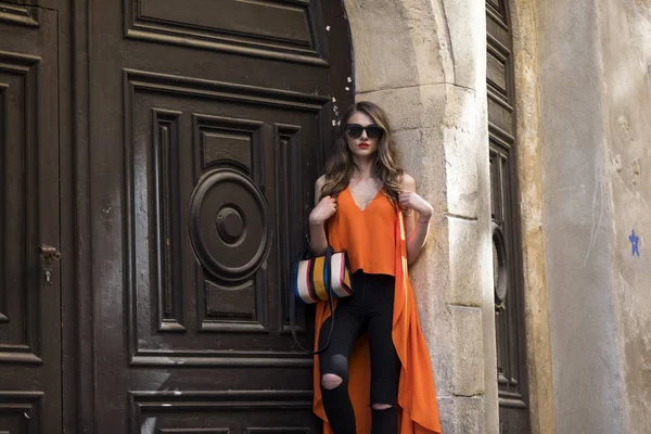 Estilo de vida retrato de uma jovem mulher elegante vestindo calças pretas, top laranja e um saco de mão listrado — Fotografia de Stock