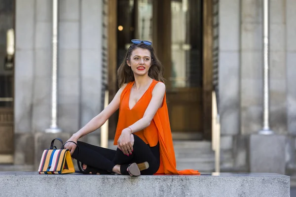 Estilo de vida retrato de uma jovem mulher elegante vestindo calças pretas, top laranja e um saco de mão listrado — Fotografia de Stock