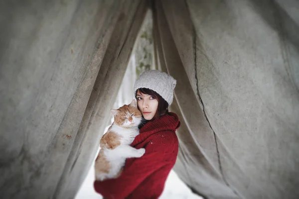 Beautiful girl with in red sweater and hat holding and playing with little fluffy cat in winter snowy park. Pets, comfort, christmas, winter and people concept young woman with cat standing outdoor.