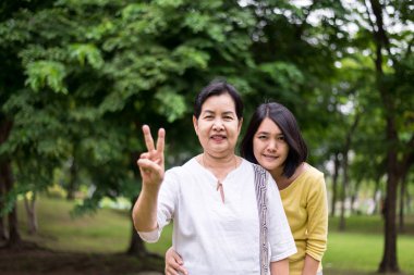 Kendine iyi bak ve destek ol, orta yaşlı Asyalı kadının kızı ile birlikte parkta iki parmağını gösteriyor, mutlu ve gülümsüyor, pozitif düşünüyor.