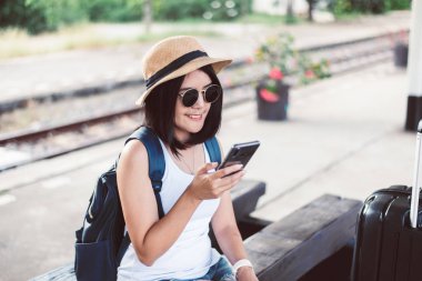 Seyahat ve tatil konsepti, mutlu Asyalı kadın turist cep telefonu kullanıyor ve tren istasyonunda bekliyor.