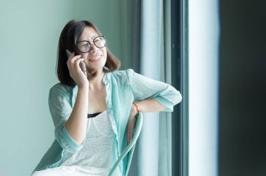 Güzel Asyalı kadın portresi rahatla ve oturma odasının penceresinin yanındaki akıllı telefonu kullan, olumlu düşün, iyi tavır, mutlu ve gülümse.