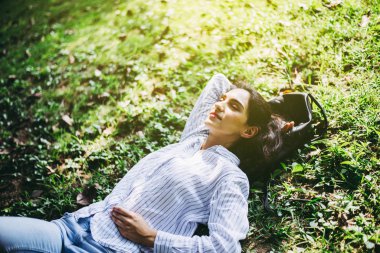 Güzel genç bir kadın ellerini başının üstüne koyup halka açık bir parkta uyuyor, dinleniyor, mutlu ve gülümsüyor.