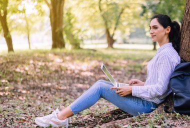 Sabahları halka açık parkta dizüstü bilgisayar kullanan güzel genç bir kadın mutlu, gülümseyen ve dinlenen bir zaman.