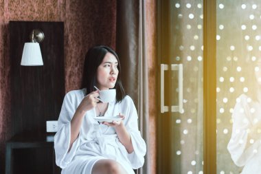 Güzel Asyalı kadın sabah kalktıktan sonra yatak odasında sıcak kahve içiyor, dinleniyor, mutlu ve gülümsüyor, sağlıklı ve hayat tarzı kavramları var.