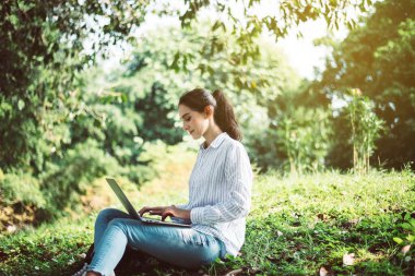 Sabahları halka açık parkta dizüstü bilgisayar kullanan güzel genç bir kadın mutlu, gülümseyen ve dinlenen bir zaman.