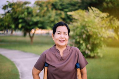 Koltuk değneği kullanan yaşlı Asyalı kadın sabah parkta yürümek için kırık bacakları destekliyor. Fizik tedavi kavramı. 